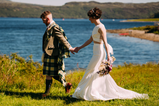 Bride and groom hold hands walking along the waterside